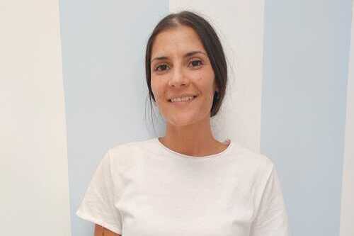 Donna sorridente con capelli castani raccolti indossa una maglietta bianca davanti a una parete con strisce verticali azzurre.