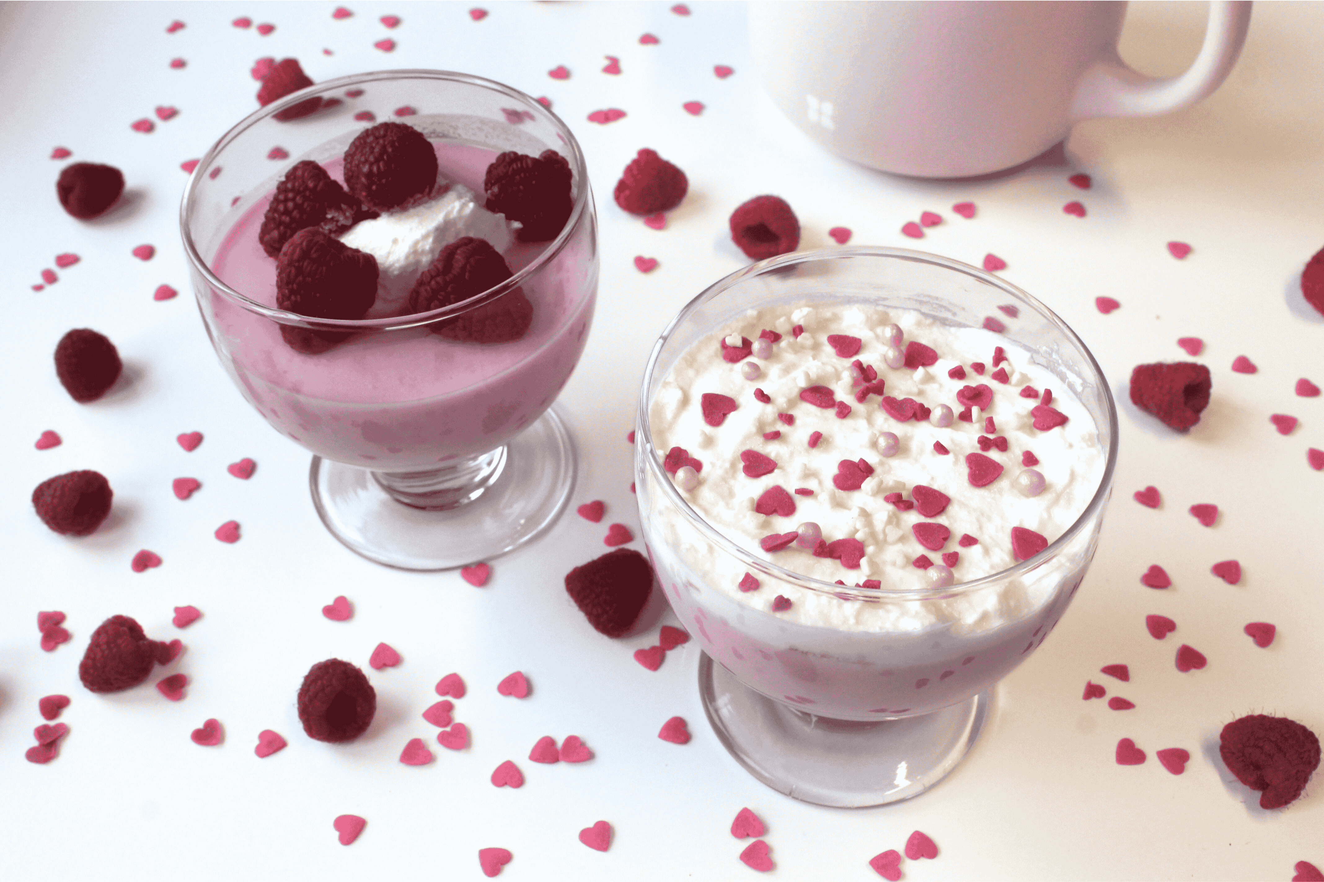 Two glass dessert cups: one with pink Greek Yogurt, raspberries, and cream, the other with yogurt, cottage cheese, and pink heart sprinkles—surrounded by raspberries and heart-shaped sprinkles for a healthy dessert on a white surface.