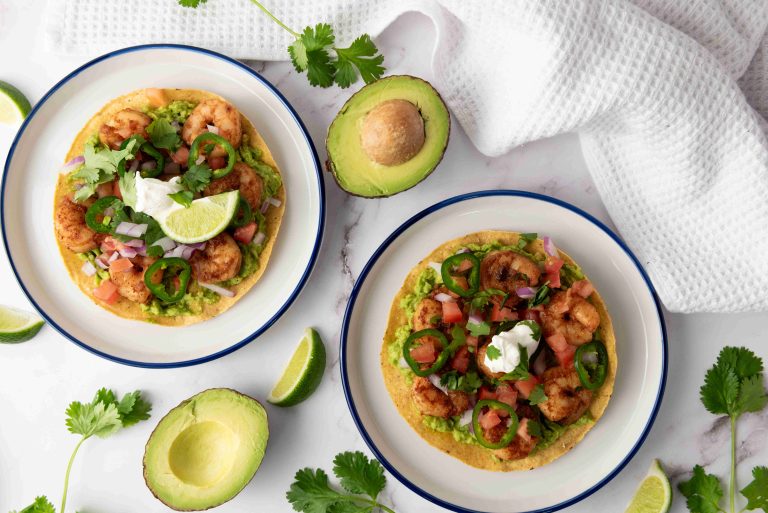 Two plates with shrimp tostadas make for a vibrant Mexican dinner, topped with guacamole, pico de gallo, jalapeño slices, sour cream, and lime wedges. Fresh avocado mash, cilantro, and lime slices are scattered on a white surface nearby.