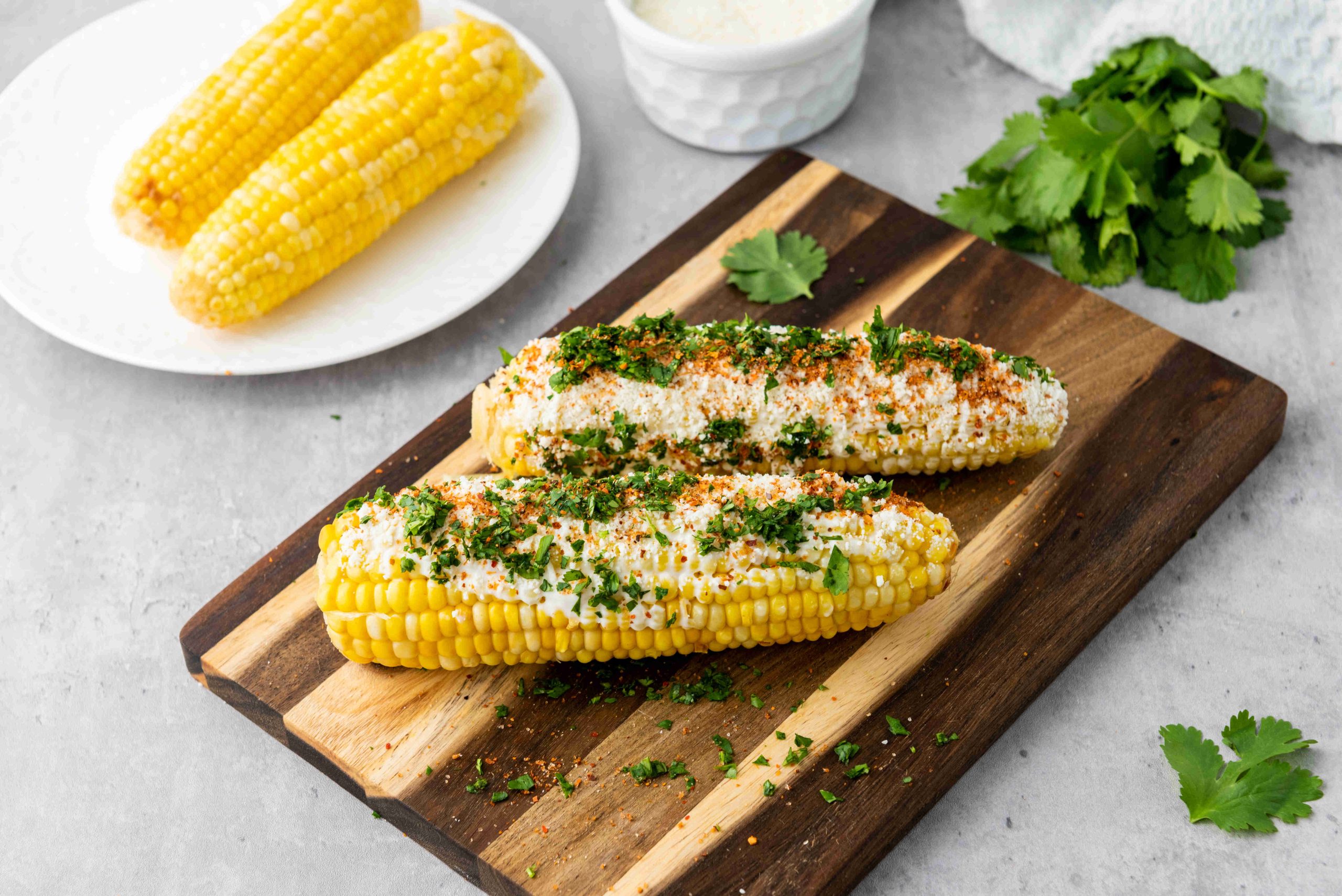 Two ears of Mexican Street Corn topped with cheese, chili powder, and chopped cilantro sit on a wooden board. More plain corn and a bowl of cheese are in the background, making this a quick Elote recipe scene with fresh cilantro on the counter.