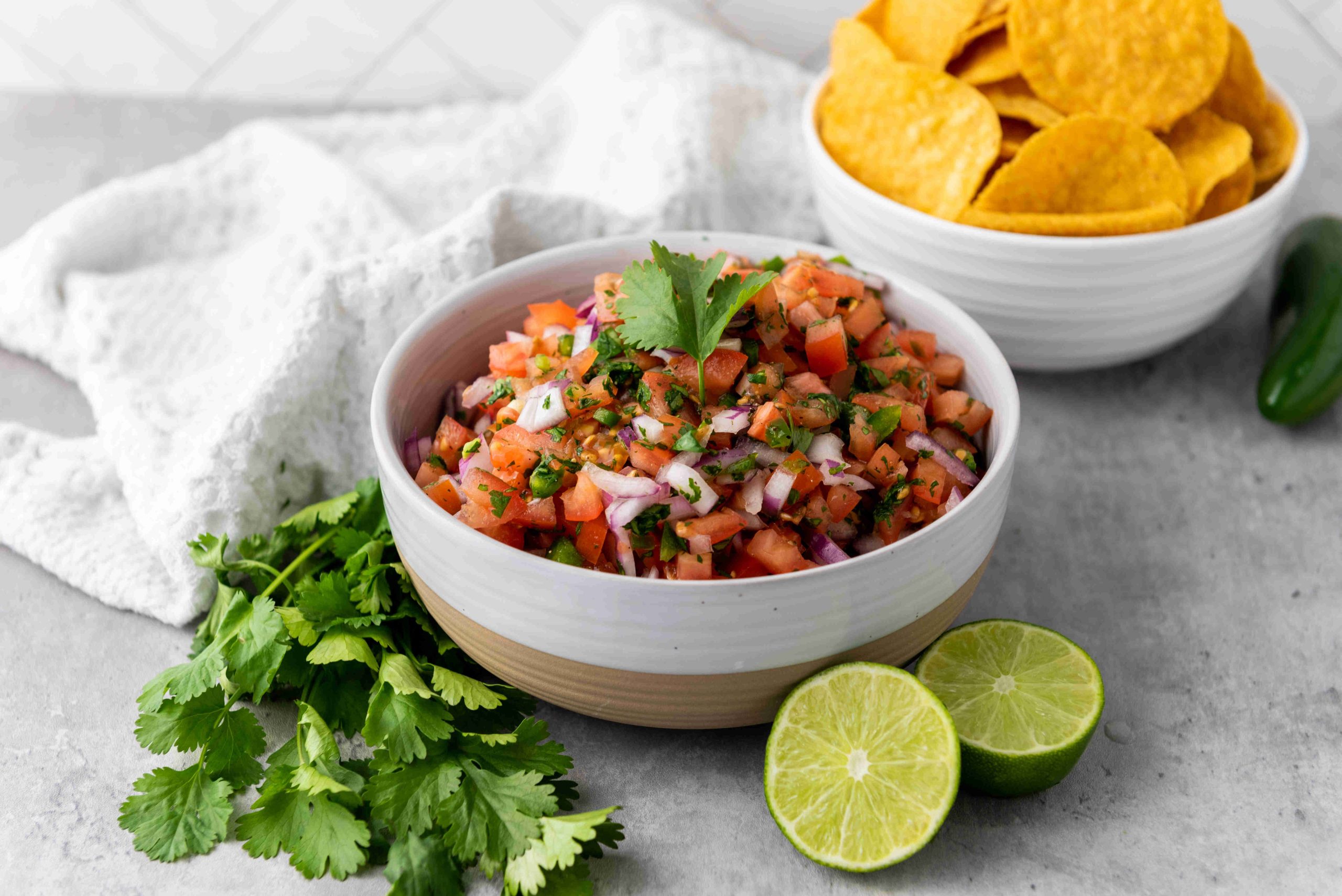 A bowl of fresh pico de gallo made with juicy fresh tomatoes sits on a gray surface, surrounded by lime halves, cilantro sprigs, a green chili, and a bowl of yellow tortilla chips. A white towel is in the background. Perfect for any homemade salsa recipe!.