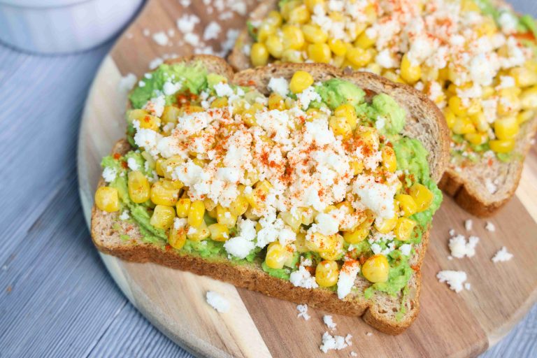 Two slices of whole grain Mexican avocado toast topped with mashed avocado, roasted corn, queso fresco, and a sprinkle of red seasoning, served on a wooden board.