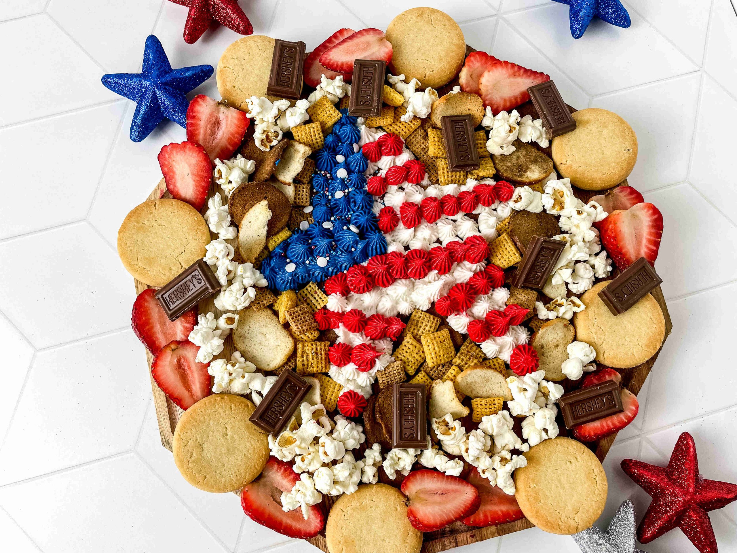 A patriotic party platter features a large star decorated with red, white, and blue icing, surrounded by cookies, chocolate, popcorn, cereal, and sliced strawberries. Red, blue, and silver glitter stars decorate the background for a festive 4th of July.