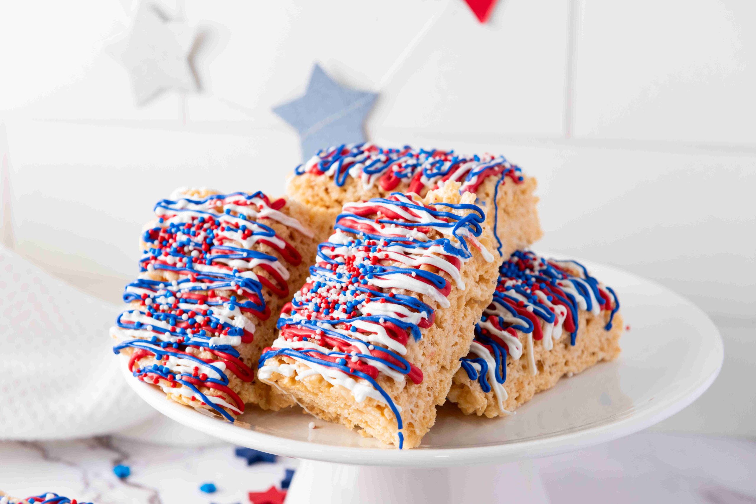 Rice Krispies treats stacked on a white cake stand, decorated with red, white, and blue icing plus festive star-shaped sprinkles—perfect 4th of July treats. Patriotic-themed star decorations hang in the background.