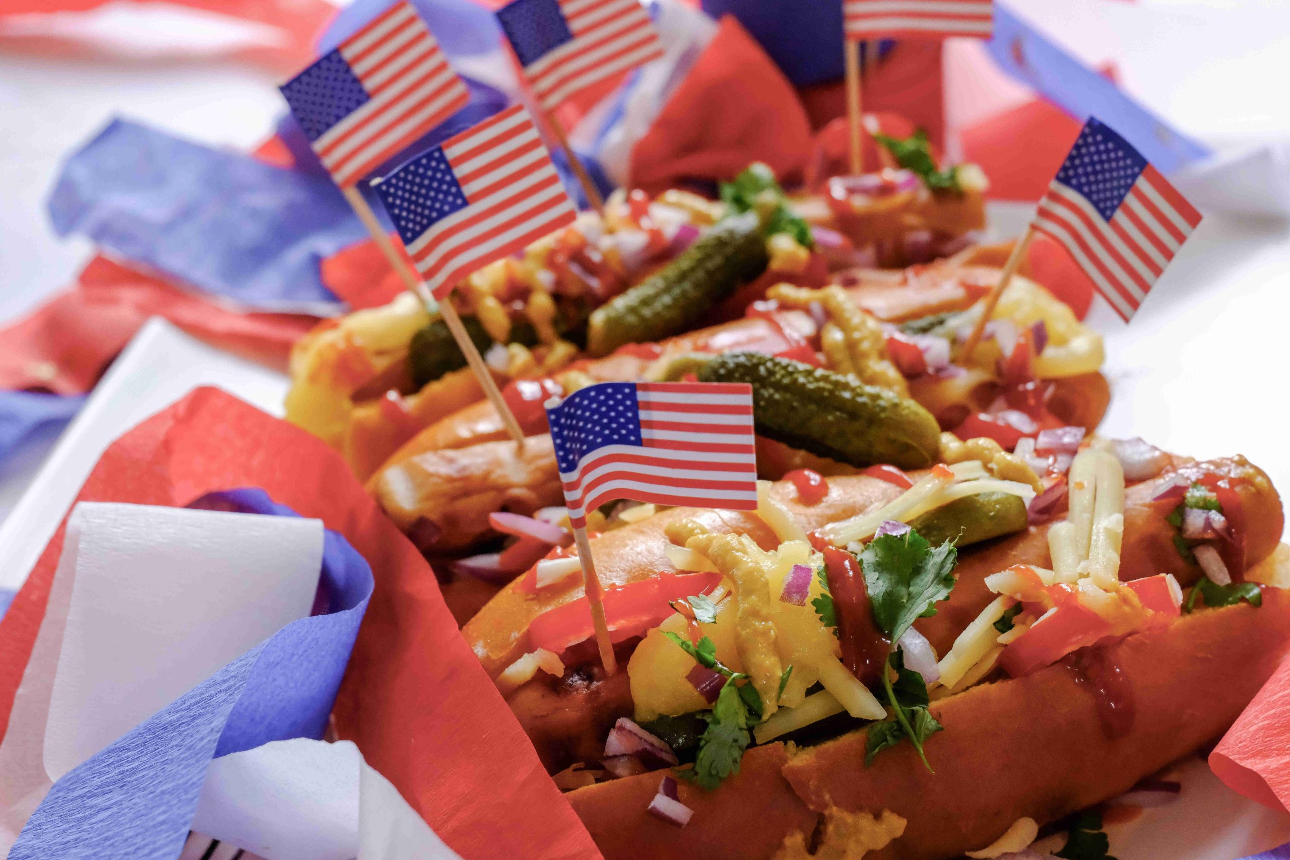 Close-up of American Hot Dogs topped with pickles, onions, cheese, and herbs, each with a small American flag. Red, white, and blue napkins add a festive touch, perfect hot dogs for a patriotic celebration.