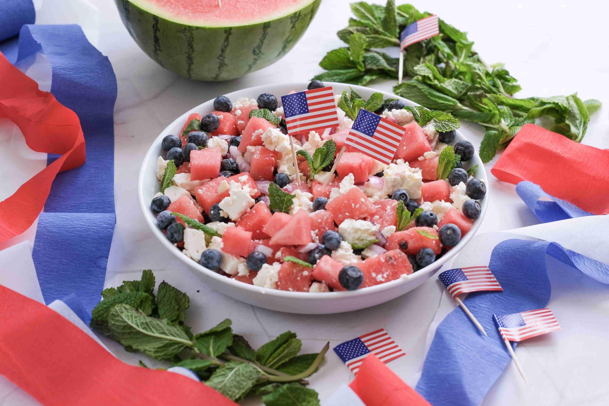 A festive Watermelon Blueberry Feta Salad in a bowl, garnished with mint and tiny American flags, is surrounded by red, white, and blue streamers—perfect for a Red White Blue Salad at your next Summer BBQ. Halved watermelon sits in the background.