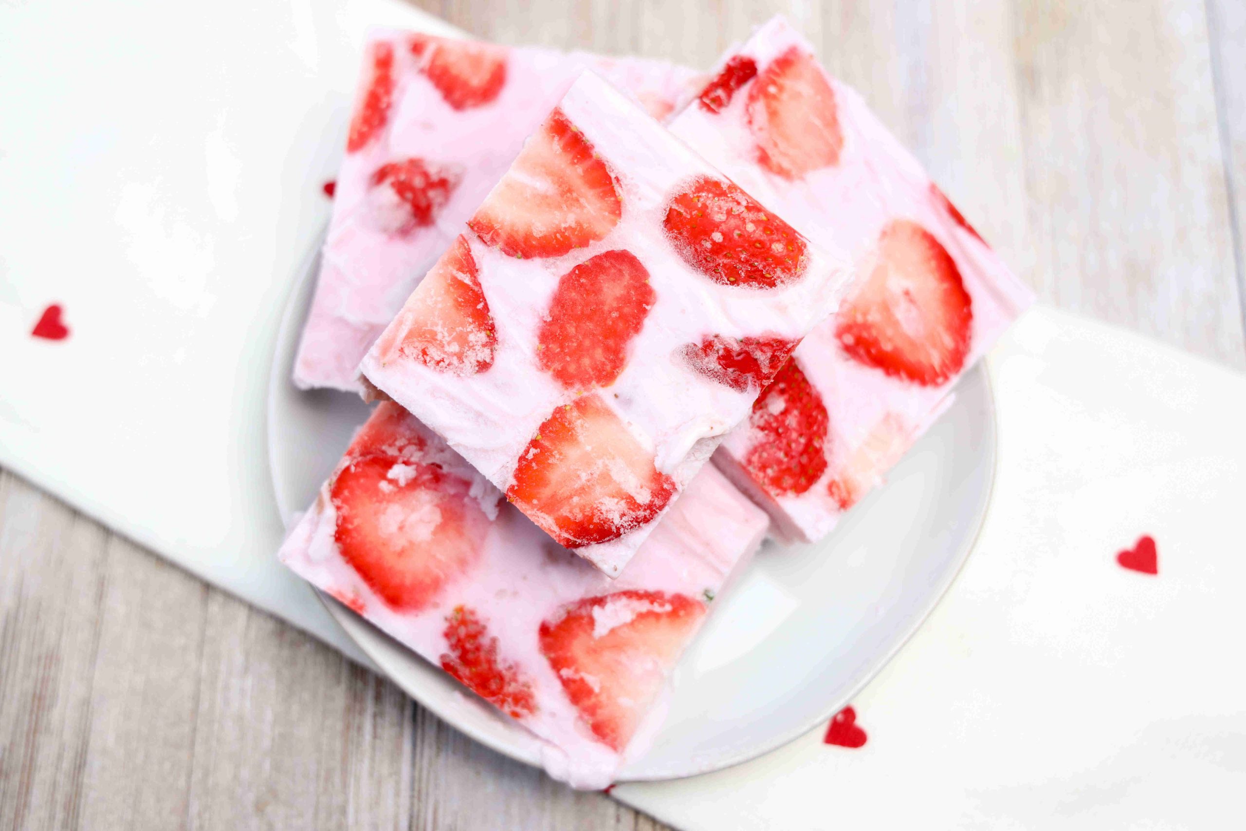 A plate stacked with pink strawberry yogurt bark bars topped with fresh strawberry slices sits on a white cloth decorated with small red hearts. The bars have a creamy texture and a fruity appearance.
