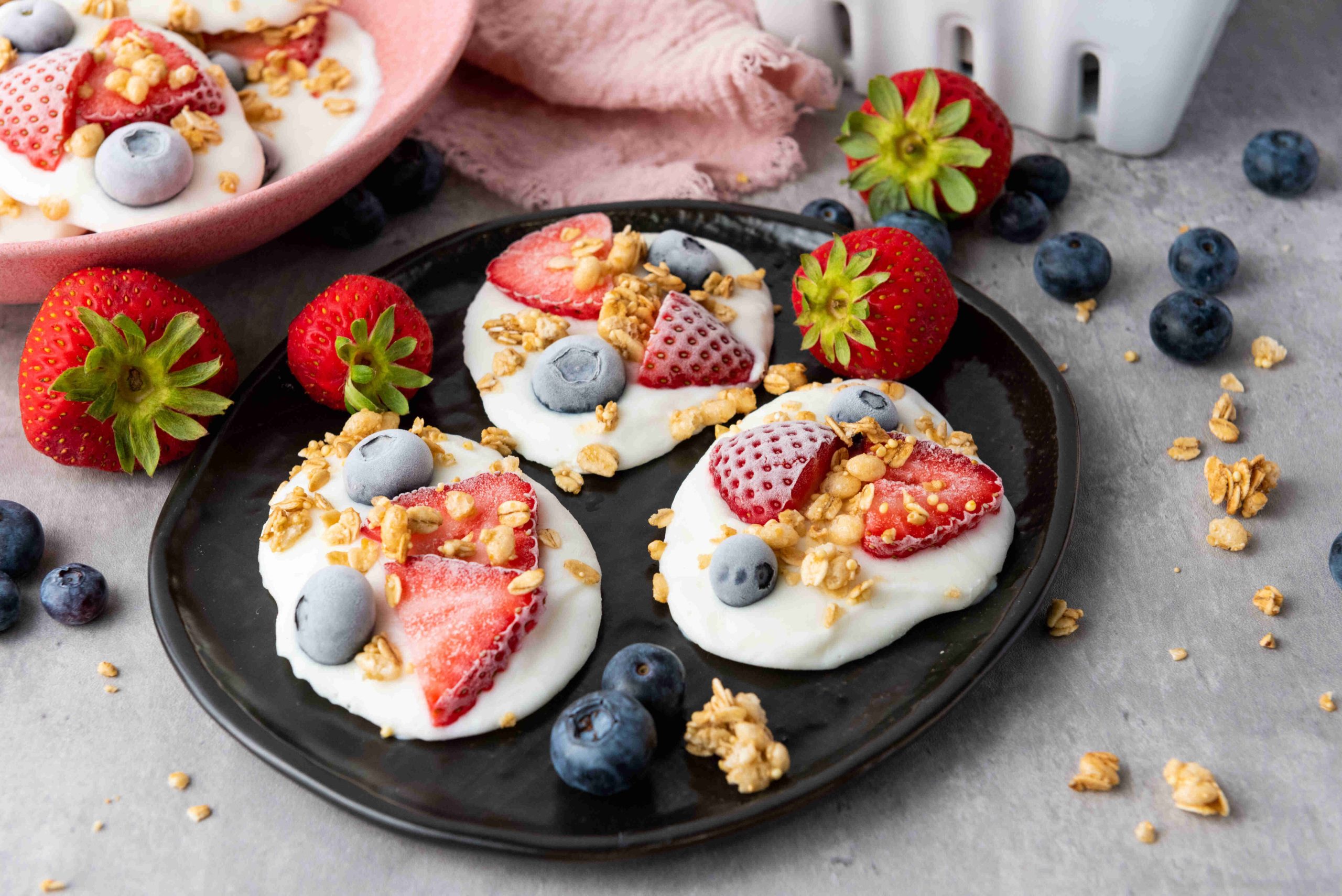 A black plate with frozen yogurt bites topped with strawberries, blueberries, and granola sits on a gray surface. Fresh strawberries, blueberries, and granola are scattered around the plate.