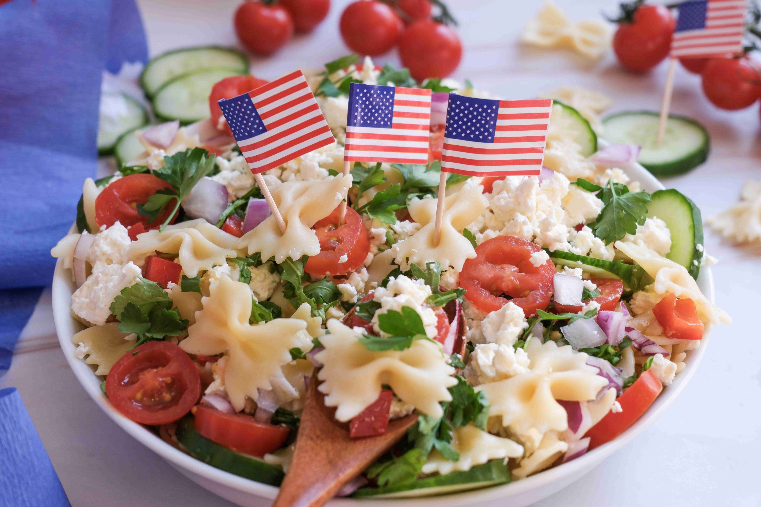 A festive 4th of July pasta salad with bowtie pasta, cherry tomatoes, cucumber, red onion, feta cheese, and parsley is topped with small American flags—a perfect red white blue side dish for your celebration. A wooden spoon rests in the bowl.