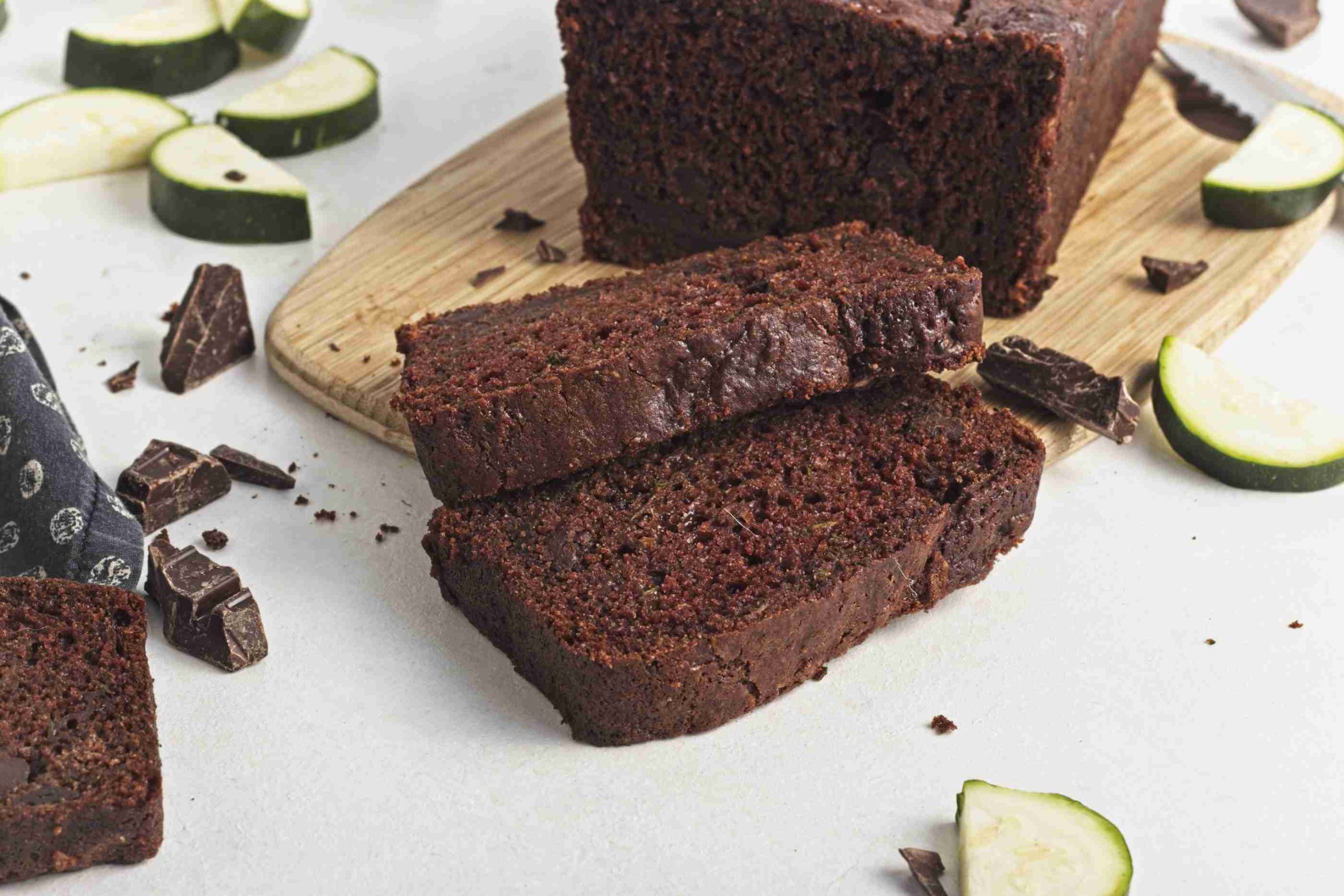 Two slices of rich vegan chocolate chip zucchini bread lie on a light surface in front of a whole loaf on a wooden board, surrounded by zucchini pieces and chunks of chocolate.