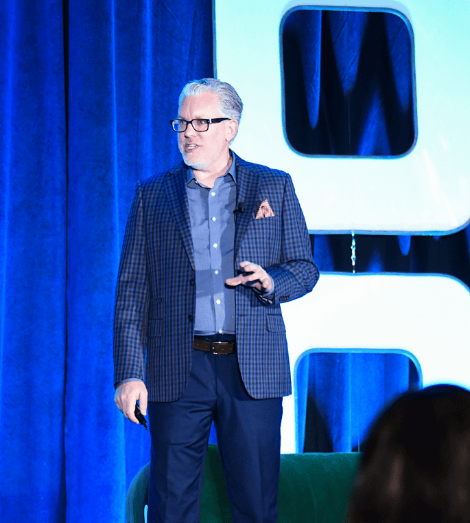 Chris Hood AI Keynote Speaker, speaking at a conference in Dallas, dressed in a checked blazer and glasses, delivering a presentation on stage with a modern backdrop.