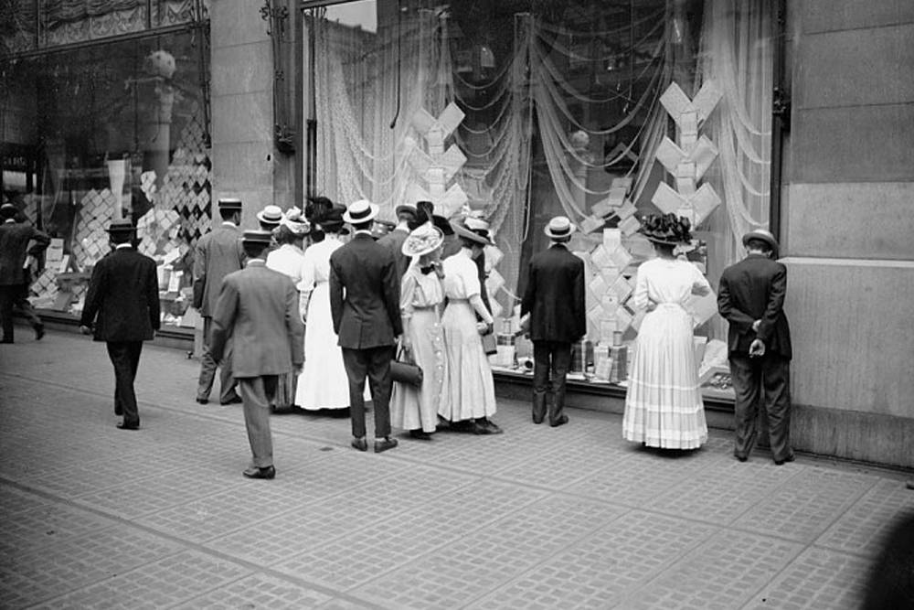 Vintage storefront window shopping with well-dressed men and women in early 20th-century attire, viewing display of books and decorative items, capturing historic urban retail scene.