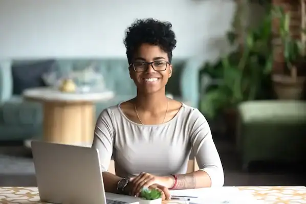 Young woman with short curly hair and glasses working at a laptop in a cozy, modern cafe setting.