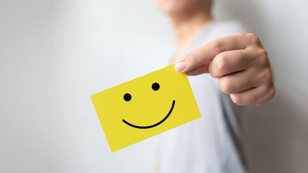 Bright yellow smiley face sticker held by a person's hand for positivity and happiness, emphasizing friendly customer service or positive attitude.