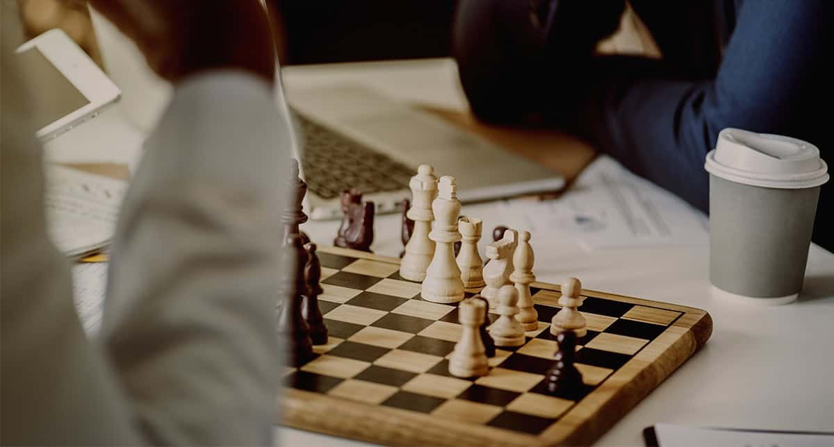 Professional chess game in progress featuring classic wooden chessboard and pieces, with players engaged in strategic gameplay during a competitive tournament or casual setting.