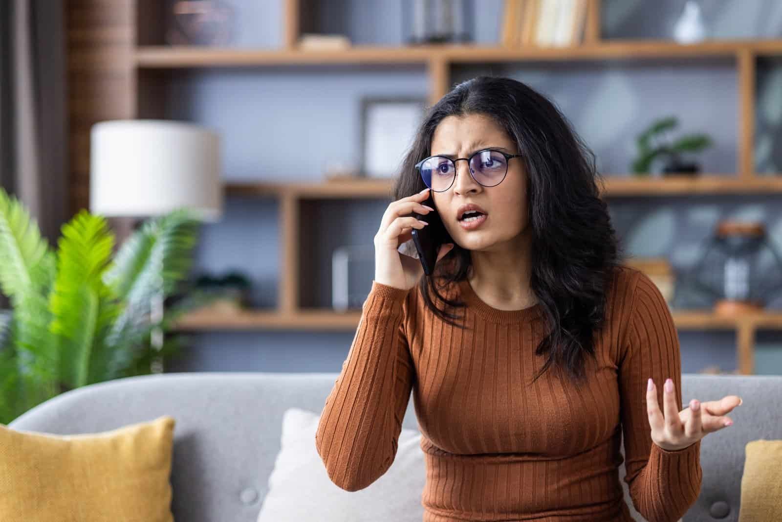 Worried young indian woman wearing glasses sitting on sofa at home and talking on mobile phone emotionally gesturing with hands » Chris Hood Unhappy customer churn