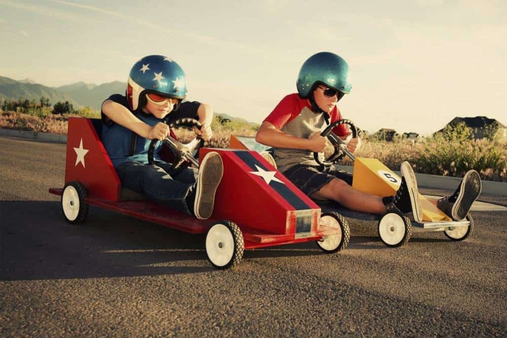 Kids racing mini cars with patriotic American flags and helmets on an open road during sunset, showcasing fun and adrenaline-filled outdoor activities.