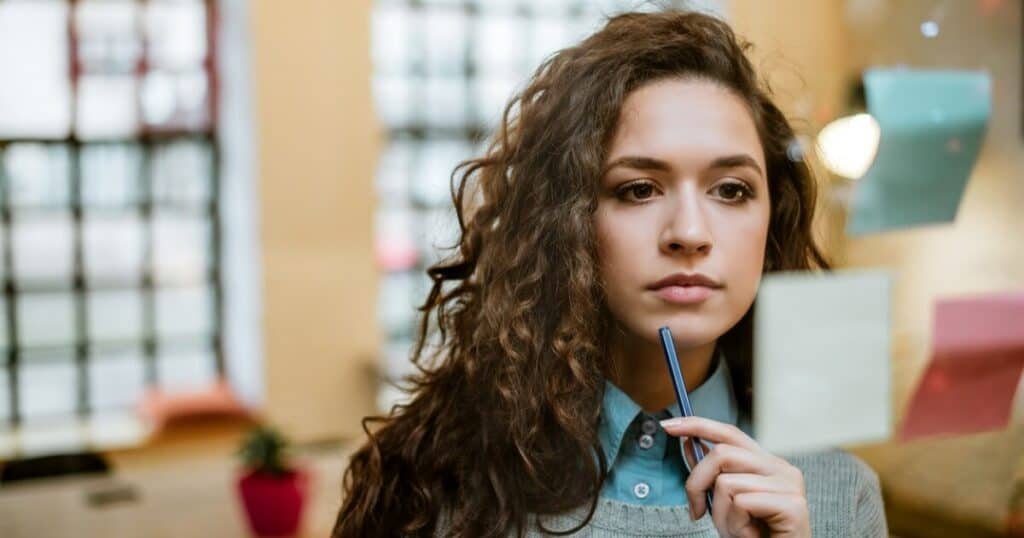 Thoughtful young woman pondering ideas in modern office environment with sticky notes on glass wall, representing creativity and strategic planning.