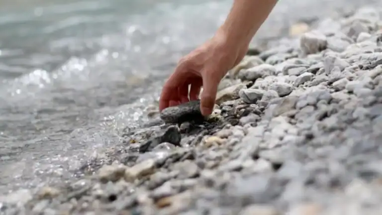 A male hand picking up a round, smooth stone near a lake symbolizing facing your giants