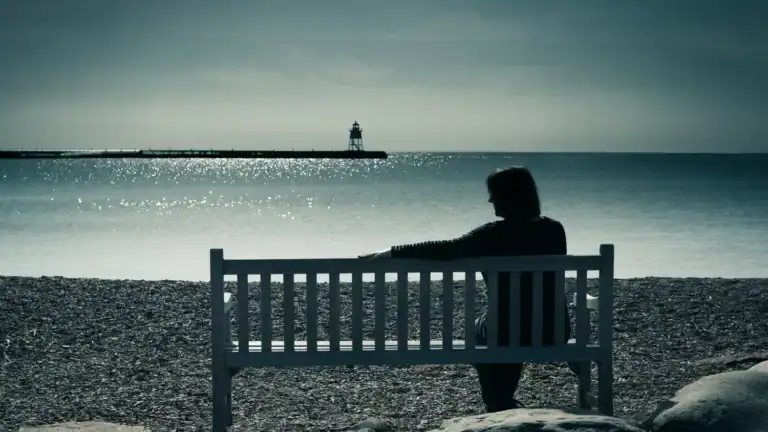 A woman sitting on a bench in front of the ocean looking at the empty seat beside her.