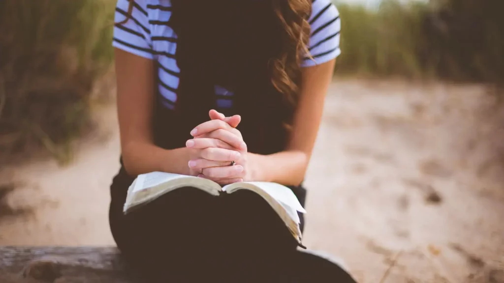 A Christian woman praying with her hands clasped on a Bible; strengthening her daily prayer.