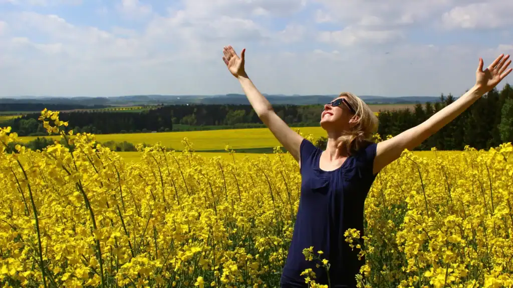 A woman standing in a field of yellow flowers with her hands in the air, facing the sun