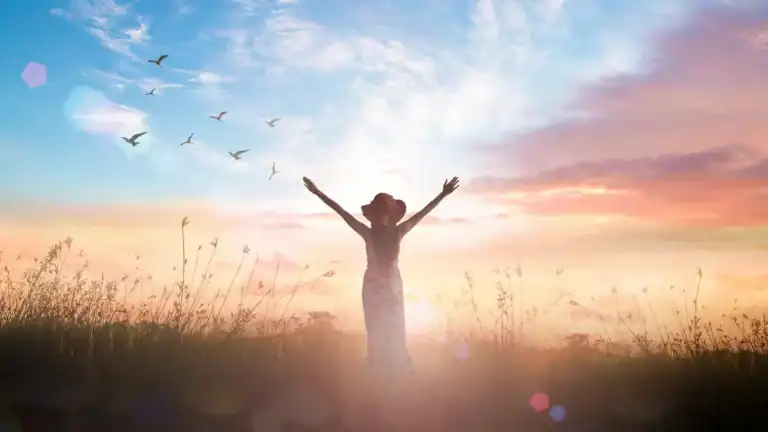 A Christian woman in field in front of a sunset with her arms raised relating to Jehovah Jireh