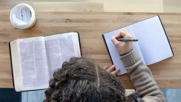 A woman sitting at a table reading a Bible and writing in a notebook symbolizing how to create a Bible study