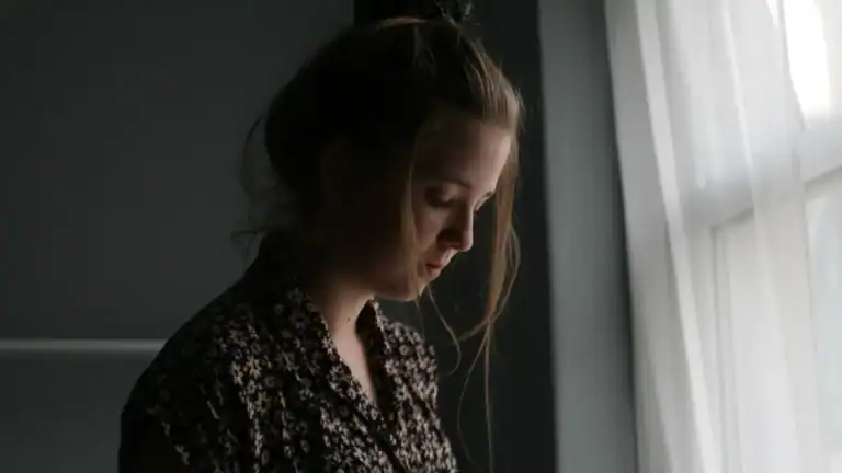 A woman standing beside a window looking down.