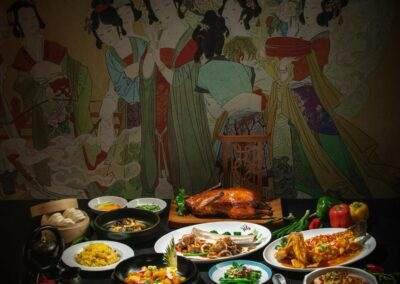 A table full of chinese food with a painting on the wall.