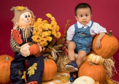 A boy in overalls sits next to a scarecrow and pumpkins.