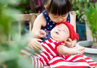 A baby in a red, white and blue outfit is sitting on a wooden bench.
