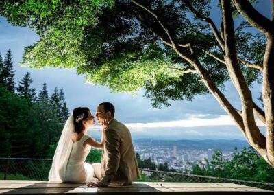 CineCrown Photography A bride and groom sitting on a bench overlooking a city.