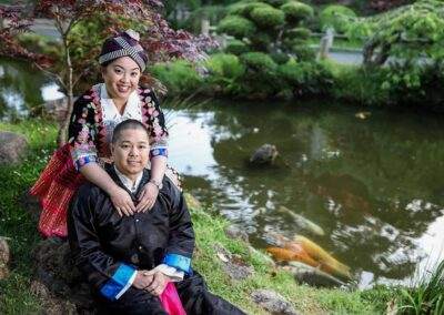 A couple in traditional hmong clothing sits on a rock next to a pond.