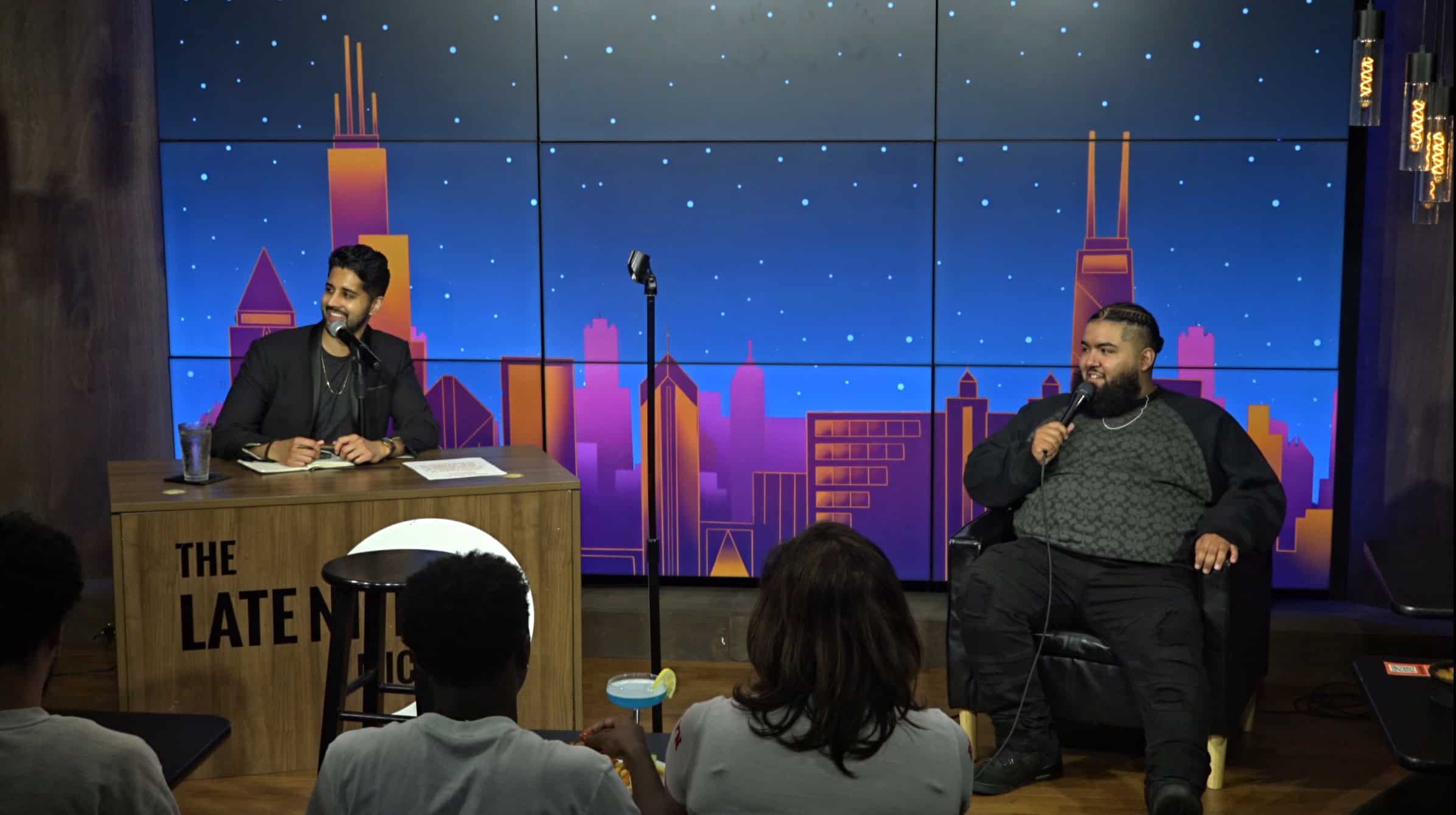 Late Nite Mic host behind desk with guest comedian on stage at Comedy Bar