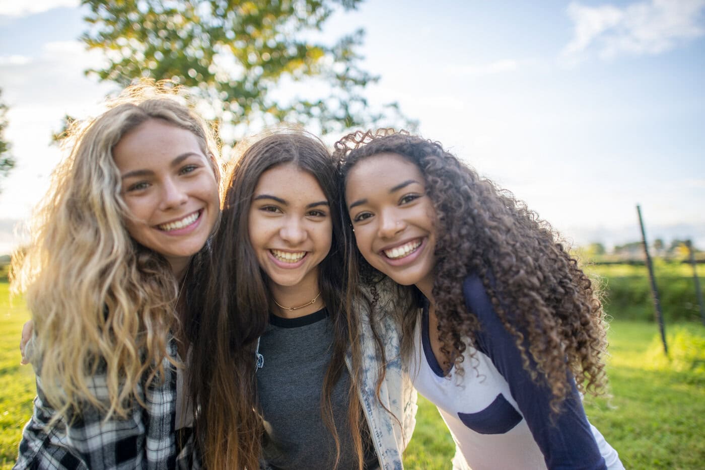 A small group of three female friends huddle in closely together as they pose for a portrait. They are each dressed casually and are smiling on the warm summers evening.