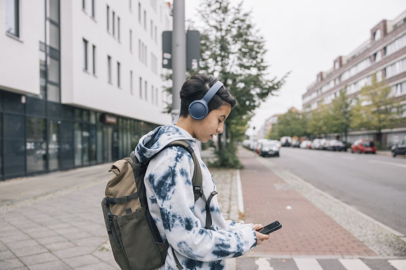 Image of teenager wearing a tie-dye hoodie, backpack, and headphones, looking at a smartphone on an urban street