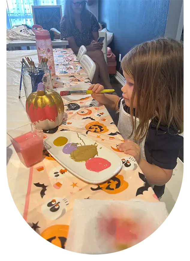 Child painting a pumpkin on a decorated table with Halloween motifs.