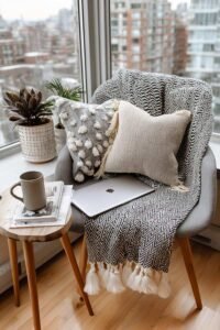 Small studio apartment corner with armchair, soft pillows, throw blanket, laptop, and city view from window
