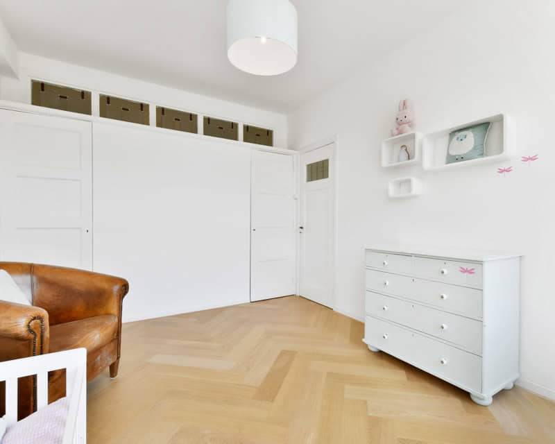 This is a bright, minimalistic nursery with white walls, a leather armchair, a white dresser, and wall shelves featuring stuffed toys and a clock. It's ready for e-design interior design by design-chat.