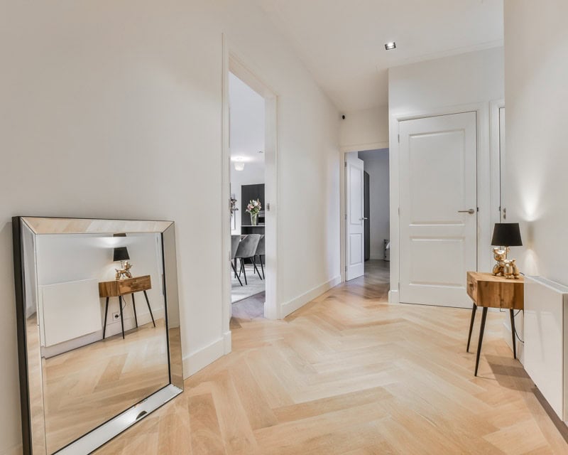An inviting hallway features a herringbone wooden floor, white walls, a large mirror, a small table with a lamp, and an open door to another room - the perfect scene for e-design interior design by design-chat.