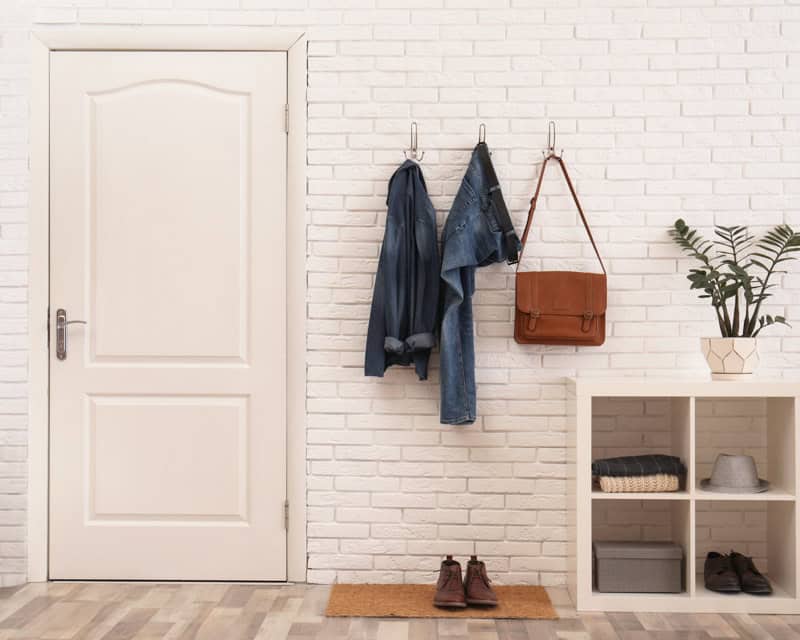 A tidy entrance hall with a closed white door, clothes hanging on hooks, a brown bag, a shoe rack, a plant, and shoes on the floor. The perfect spot for a new e-design interior design package by design-chat