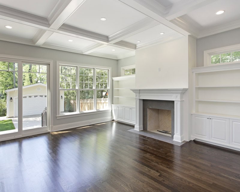 This image shows an empty, bright living room with a fireplace, built-in shelves, large windows, a dark wooden floor, and a tray ceiling in need of help with a paint palette e-design interior design from design-chat.