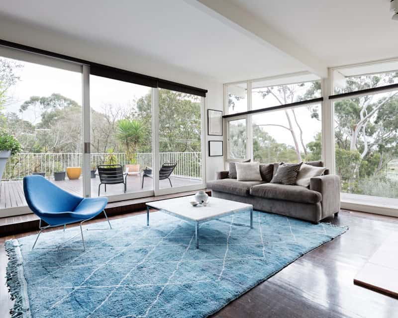 Contemporary living room with large windows, a plush sofa, blue accent chair, glass coffee table, and a view of a wooded area outside. This room is in need of a room layout e-design interior design by design-chat.