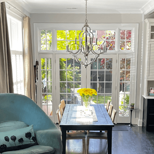 Bright, elegant dining room with large windows, a wooden table, ornate chandelier, fresh yellow flowers, and a blue armchair. Sunlight filters through, illuminating the space of this e-design interior design outcome.