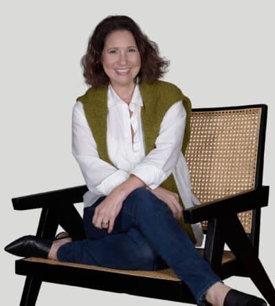 A smiling person sits casually on a black director's chair, wearing a white shirt, green vest, blue jeans, and black shoes, against a white background.
