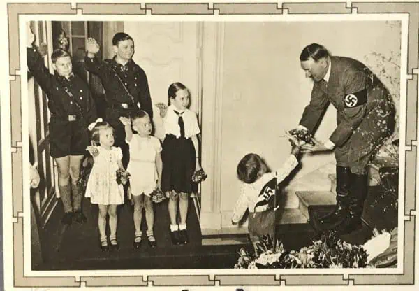 Nazi officer presenting a medal to a young boy in a uniform, with children and adults observing in t.