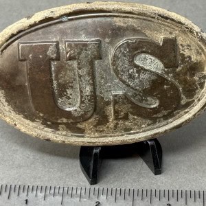 U.S. military belt buckle with embossed "U.S." letters, displayed on a small stand.