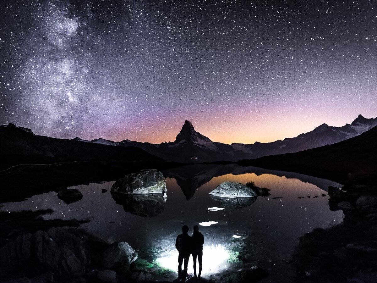 A silhouette of two people and the Matterhorn among a starlit sky and the Milky Way.