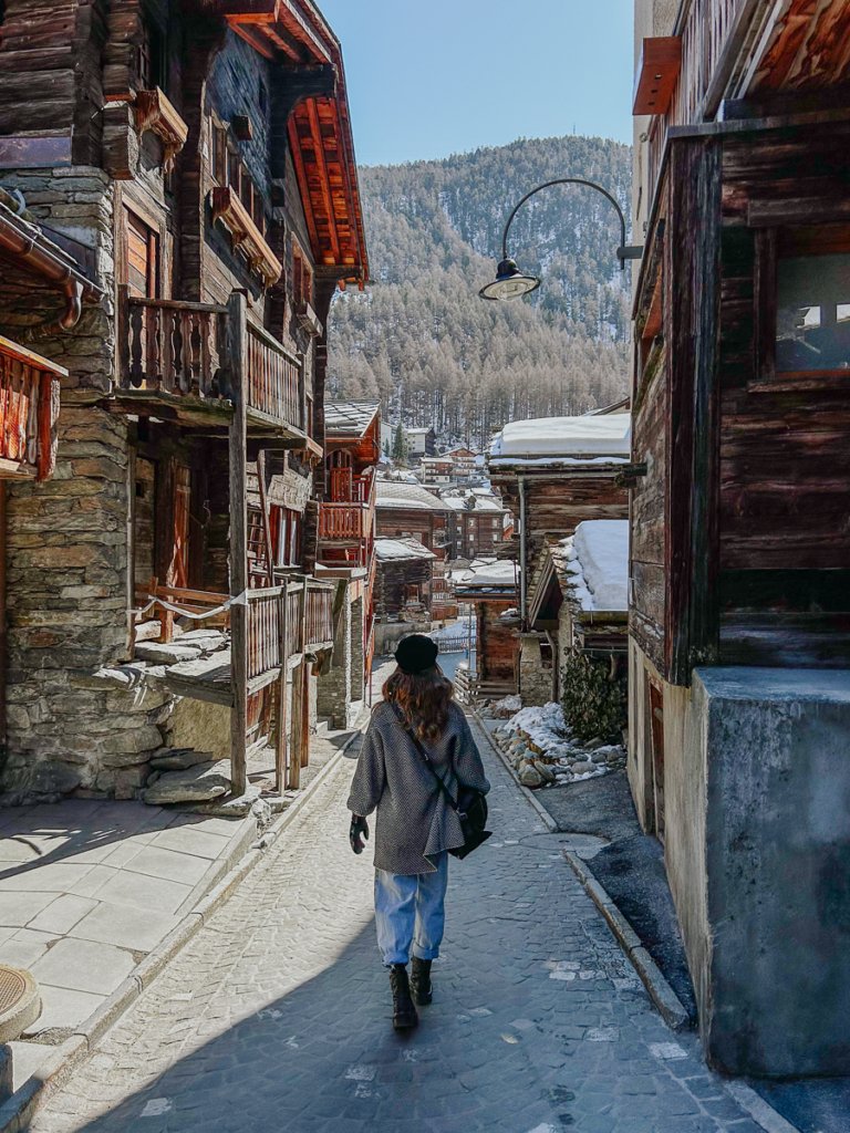 Person walking down Hinterdorfstrasse among old wooden Swiss buildings in Zermatt.